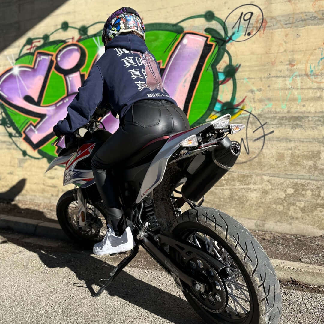 Person im INFA 295 BIKELIFE Hoodie auf einem Motorrad vor einer Graffiti-Wand, mit einzigartigem Design auf dem Rücken