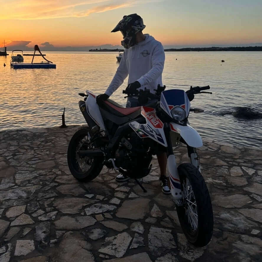 Person im INFA 295 ONE LOVE Hoodie steht mit Motorrad am Meer bei Sonnenuntergang.