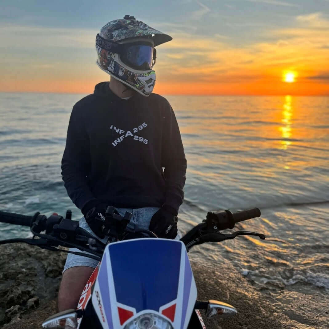 Person trägt einen INFA 295 BIKE LIFE Hoodie, sitzt auf einem Motorrad am Meer beim Sonnenuntergang.